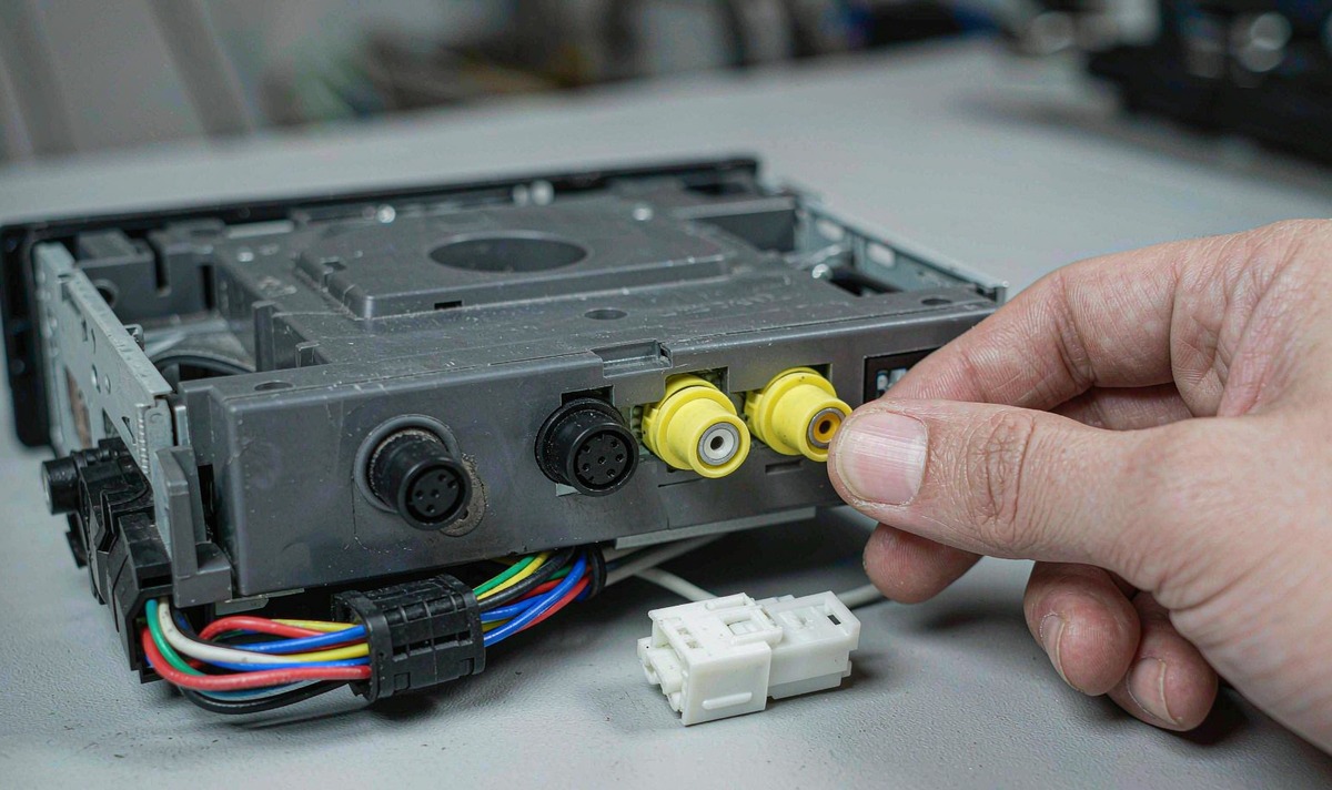 Back panel of a car stereo head unit on a workbench with yellow RCA video sockets, a multi-pin plug and a hand holding one RCA connector