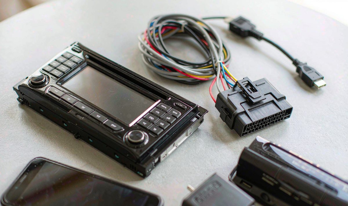 Rows of consumer video sources lined up on a workbench in front of a car screen: a phone, a laptop, a game console dock and a small streaming stick, each with its own power lead