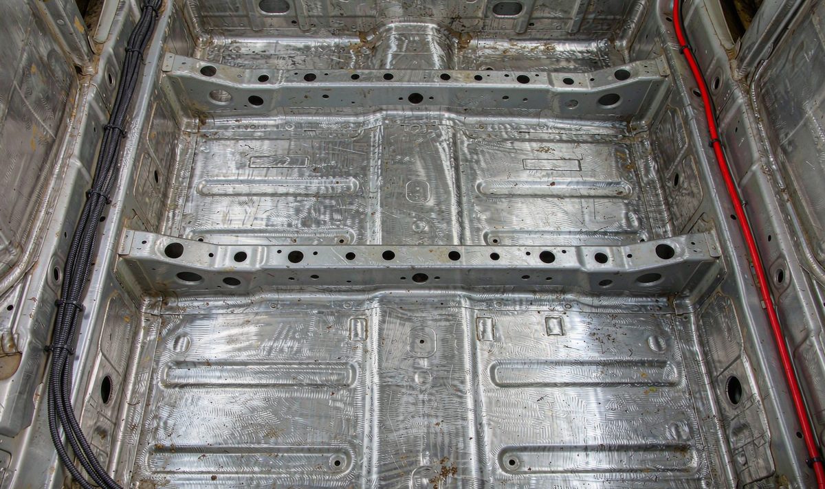 Bare metal floor pan of a car seen from above with a shielded signal cable bundle along one side and a thick red power cable along the other