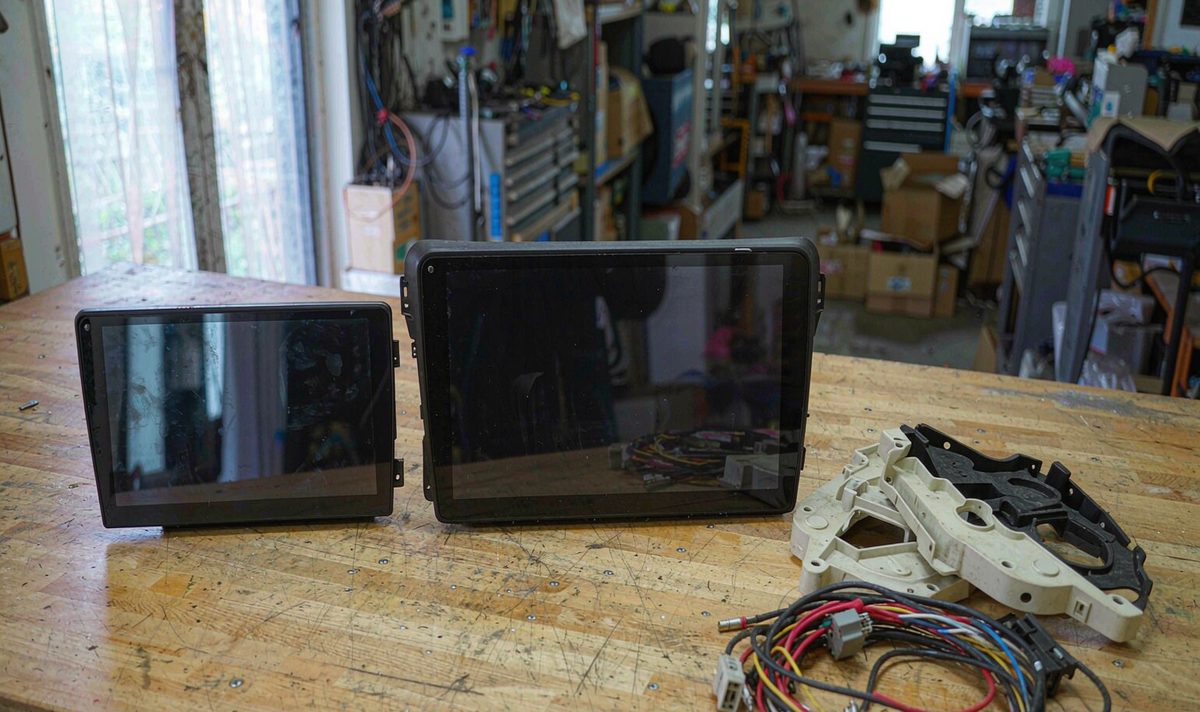 10.25 inch android head unit aftermarket dash kit panel lying beside a 12.3 inch panel on a workbench with frames and harness