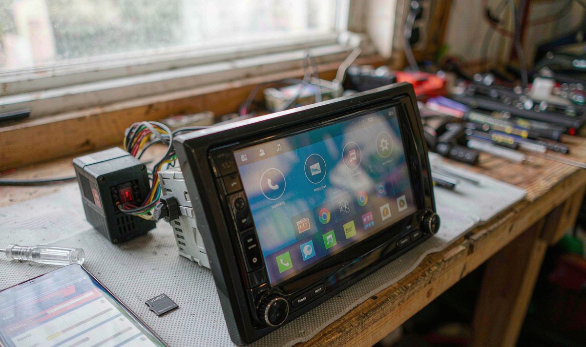 Bench test of an android head unit car stereo running a storage benchmark beside a phone showing sequential and random read results, aftermarket dash kit, retain original factory features