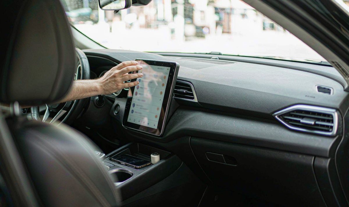 Hand reaching toward a retrofit android head unit showing a contact list, with a paired phone resting in the console, aftermarket dash kit, retain original factory features