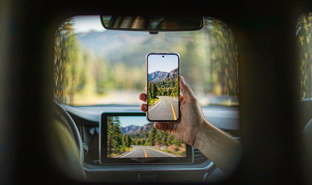 Phone in hand showing a mountain road video while the retrofit car screen below shows the same scene a moment later, car screen mirroring delay check