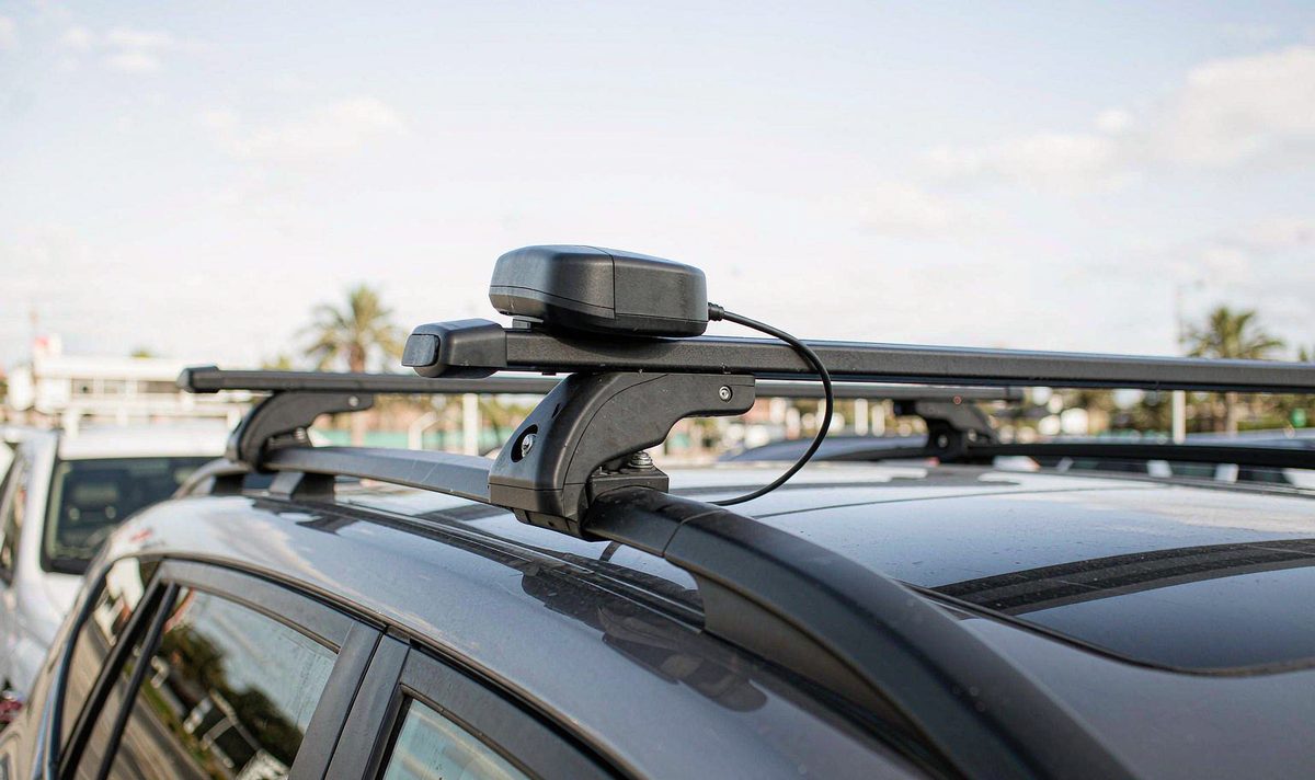 A puck mounted on or beside a crossbar loses the low satellites the fix depends on Black GPS antenna puck clamped beside a steel roof rack crossbar on a car roof, the bar casting shade over the mounting point